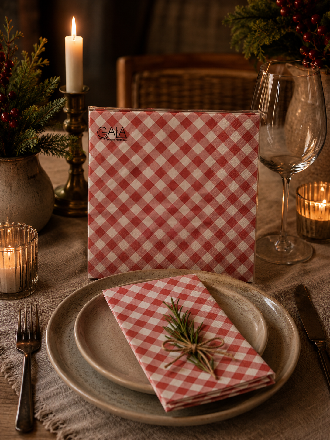 Pack of red and white gingham Gala paper napkins propped on a candlelit Christmas dinner table with a folded napkin tied with twine and fresh rosemary on a ceramic plate, wine glasses and festive greenery with red berries in the background — Mistletoe Home