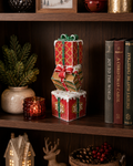 Three-tier glittery stacked gift box Christmas decoration in red, gold and green with snow detail, ribbon bows and gingerbread motifs, styled on a dark timber bookshelf with pine branches, red votive candle and Christmas books — Mistletoe Home