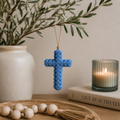 Cornflower blue floral resin hanging cross suspended from an olive branch, styled on a timber tray with wooden beads, candle and coffee table book