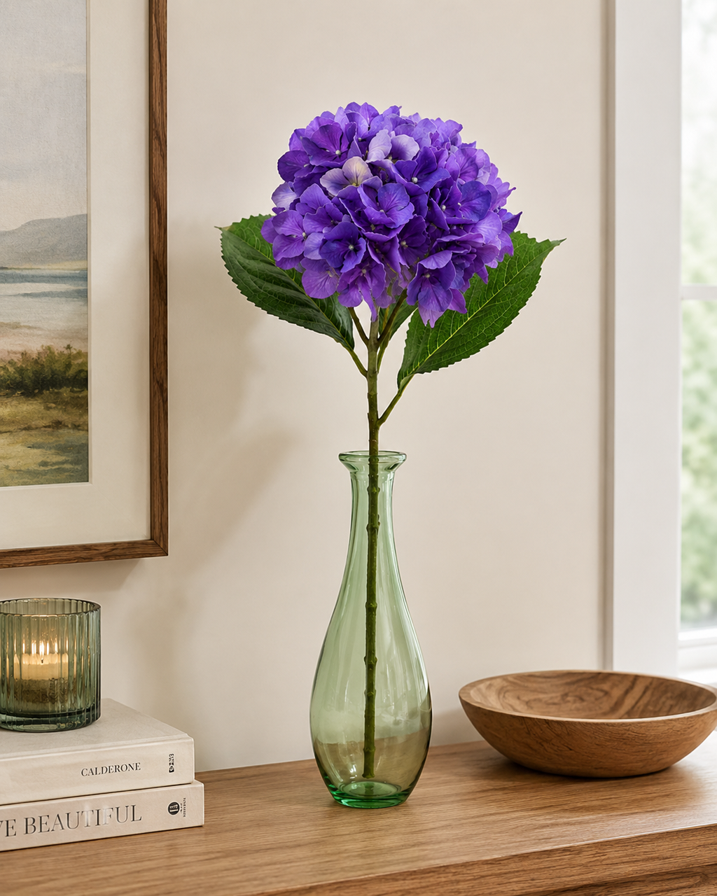 Single deep purple faux hydrangea stem in green glass vase on timber shelf with coastal artwork, candle and wooden bowl