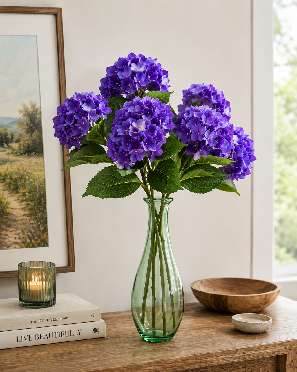 Multiple purple faux hydrangea stems arranged in a green glass vase on a timber console, styled with candle, coffee table books and landscape artwork