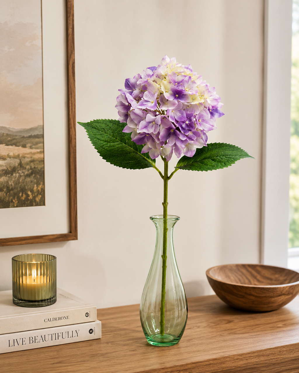  Single lilac and mauve faux hydrangea stem in green glass vase on timber shelf with landscape artwork, candle and wooden bowl