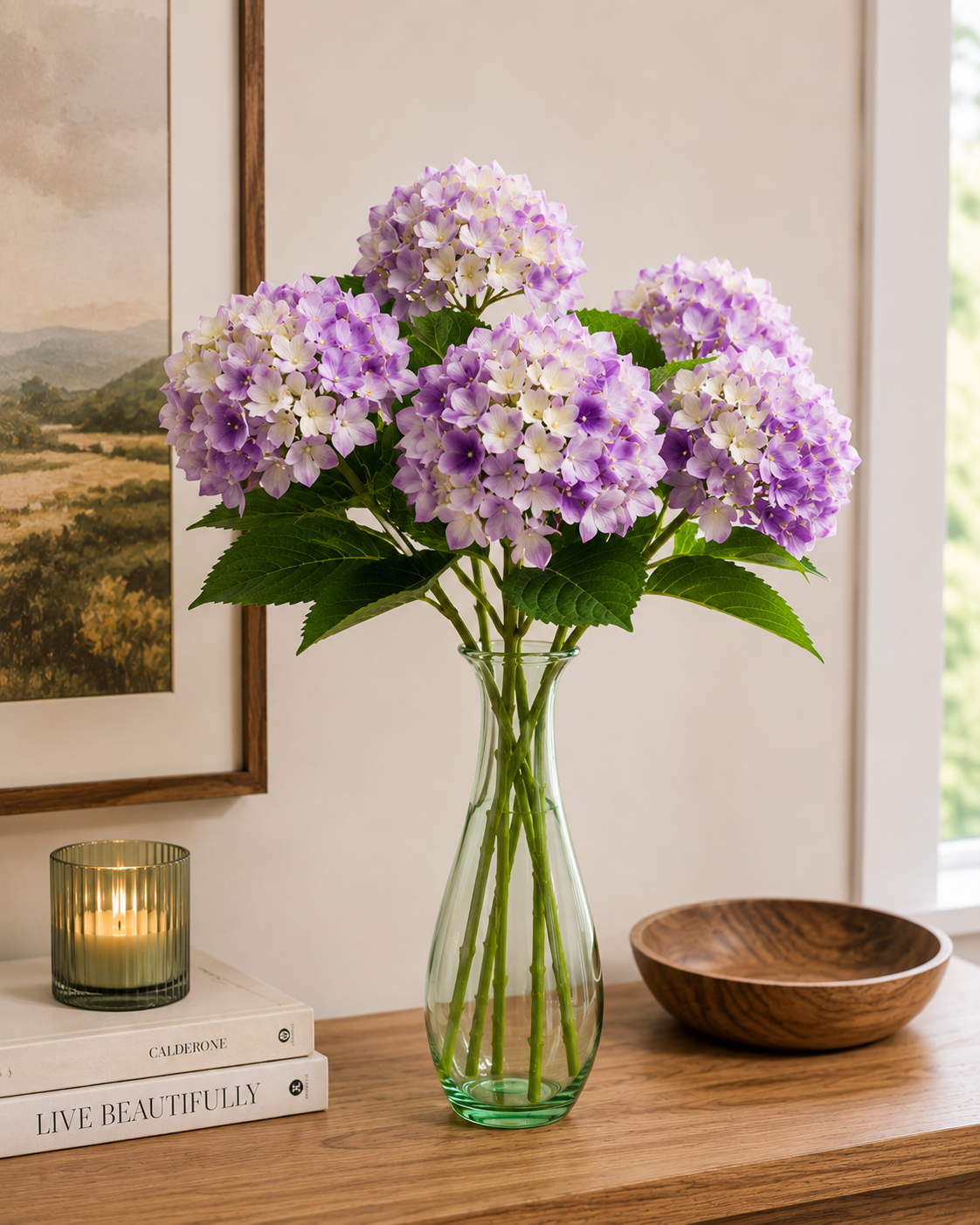 Multiple lilac faux hydrangea stems in a green glass vase on a timber console, styled with candle, coffee table books and landscape artwork