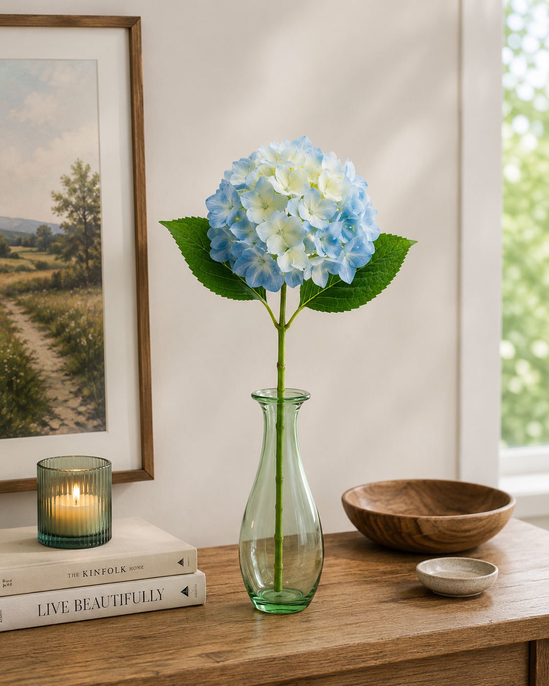 Single sky blue faux hydrangea stem in green glass vase on timber shelf with landscape artwork, candle and wooden bowl
