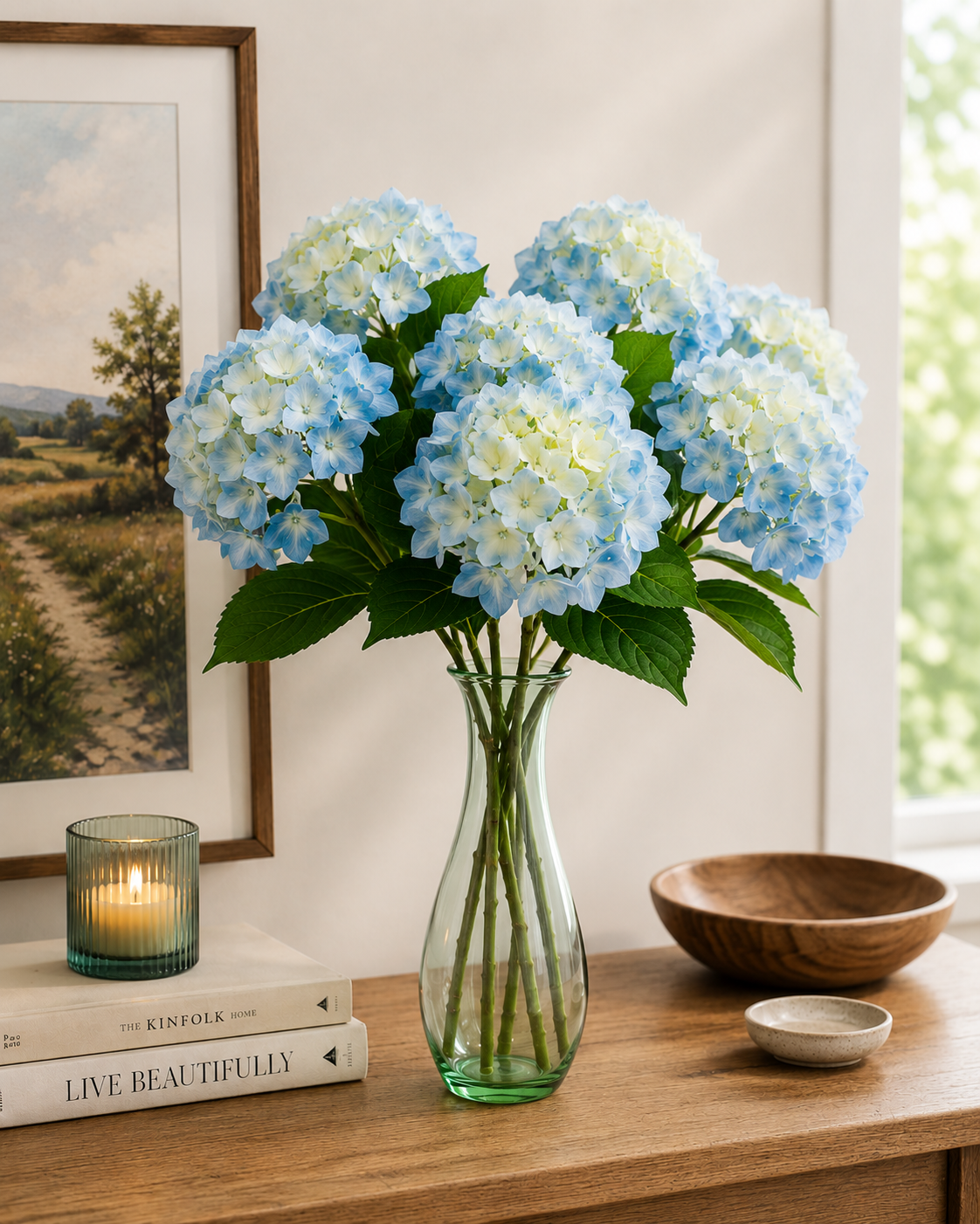 Multiple blue faux hydrangea stems arranged in a green glass vase on a timber console, styled with candle, coffee table books and landscape artwork