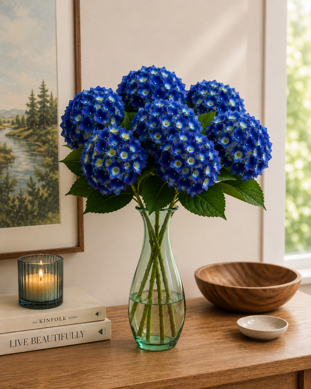 Multiple deep cobalt blue faux hydrangea stems in a green glass vase on a timber console, styled with candle, coffee table books and landscape artwork