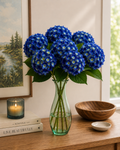 Multiple deep cobalt blue faux hydrangea stems in a green glass vase on a timber console, styled with candle, coffee table books and landscape artwork