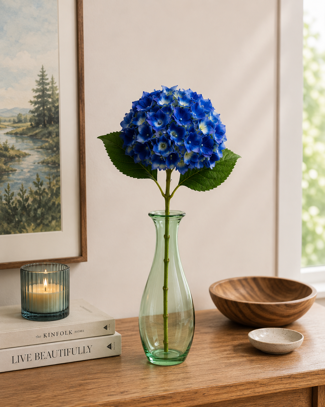 Single dark blue faux hydrangea stem in green glass vase on timber shelf with landscape artwork, candle and wooden bowl