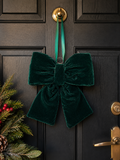 Emerald green velvet bow hanging on a dark front door next to a Christmas wreath with pine, holly and red berries