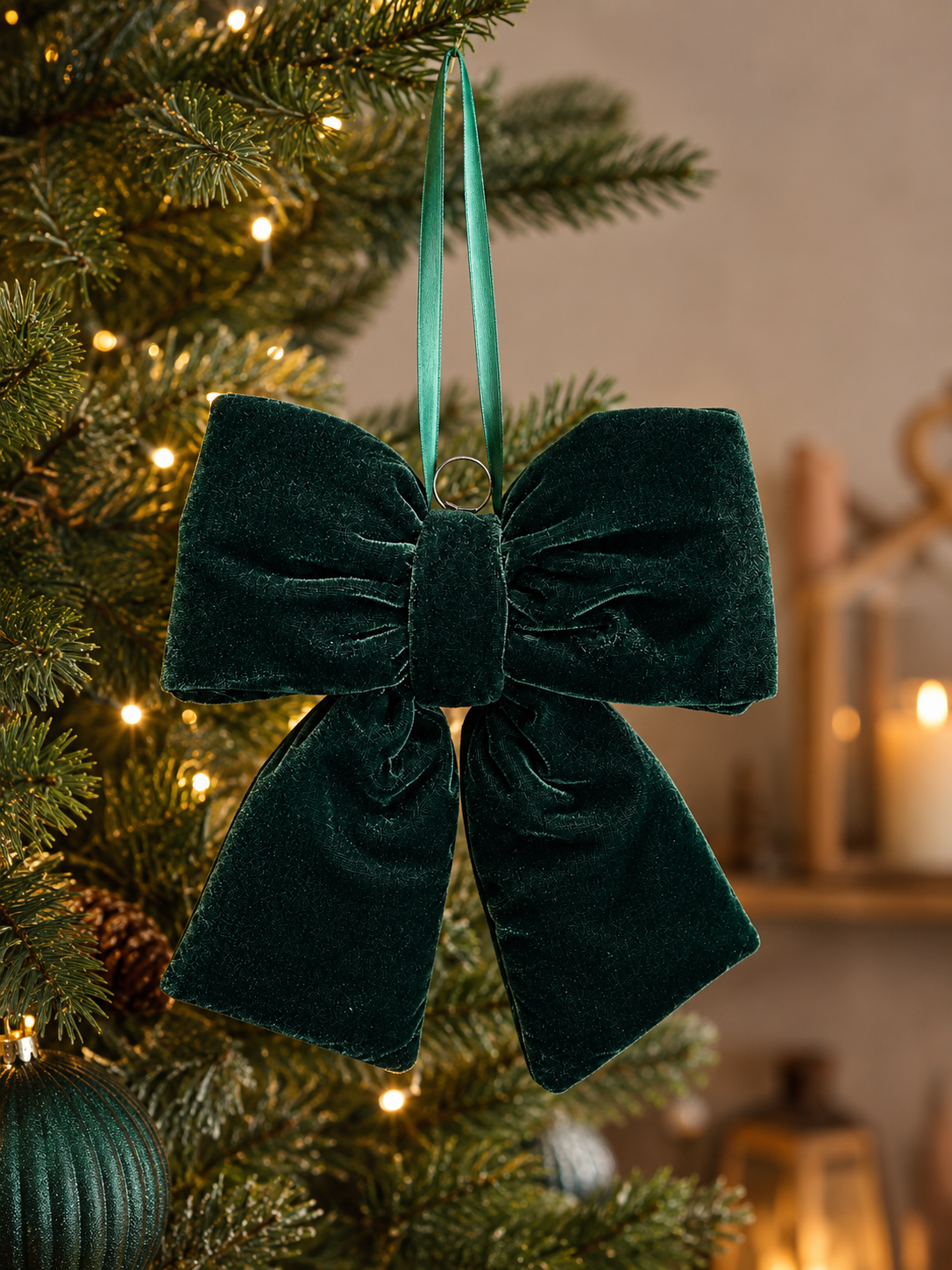 Emerald green velvet bow hanging on a lit Christmas tree with warm fairy lights and candles in the background