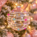 Iridescent pink glass bauble wrapped in white cord with fuchsia, gold and teal pearl beads, hanging on a frosted Christmas tree with fairy lights and pink baubles