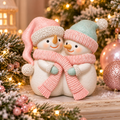 Two cuddling snowmen figurine in pink and mint hats sharing a pink knit scarf, styled on a shelf with a frosted Christmas tree, rose gold baubles and fairy lights