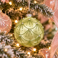 Pistachio green glass bauble with green glitter scallops and pearl beads hanging on a snowy Christmas tree among blush pink baubles and fairy lights