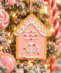 Pink iced gingerbread house ornament with white swirl roof piping and red and mint candy dots hanging on a snowy Christmas tree with pastel baubles and fairy lights