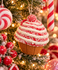Red and white swirl cupcake ornament with sparkly icing and a red cherry hanging on a snowy Christmas tree among candy cane decorations and fairy lights