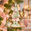Pastel mouse ornament in a pink coat and mint green earmuffs holding a small Christmas tree, hanging on a Christmas tree with pink ribbon, baubles and fairy lights