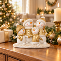Gold and white snowman family figurine with four smiling snowmen in gold hats and scarves on a wooden sideboard beside a candle, wrapped gift and Christmas tree