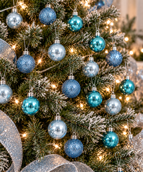 Blue and teal Christmas baubles hanging on a frosted Christmas tree with fairy lights and silver ribbon, showing gloss and glitter finishes mixed together