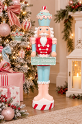 Pastel cupcake Christmas nutcracker displayed beside a flocked Christmas tree with pink and red baubles, holding a mint "Merry Christmas" sign, styled with wrapped gifts and a white lantern