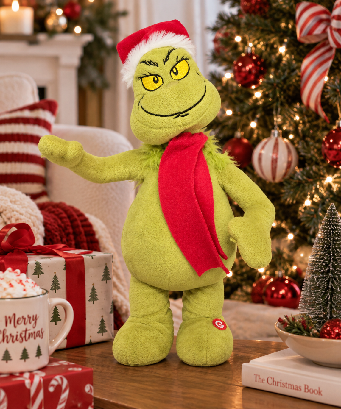 Animated Grinch plush toy wearing a red Santa hat and red scarf, standing on a wooden table in front of a decorated Christmas tree with baubles and gift boxes