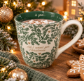 Cream ceramic mug with green sprig pattern and "All I want for Christmas is you" text, filled with hot chocolate and marshmallows, styled with Christmas lights and gingerbread