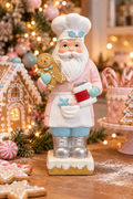 Pastel Baker Santa figurine in a pink coat holding a gingerbread man and cookies jar, styled beside a gingerbread house with warm fairy lights in the background