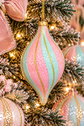 Pink and mint glitter finial ornament with gold swirl stripes hanging on a pastel Christmas tree, pink velvet bow and warm fairy lights in the background
