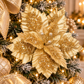 Metallic gold glitter poinsettia flower stem nestled in a gold Christmas tree, gold ribbon and warm fairy lights in the background