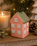 Pink and sage green ceramic house Christmas ornament displayed on a wooden shelf near a lit candle, pinecone, frosted foliage and a white ceramic tree.