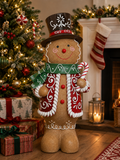 Large gingerbread man statue in a top hat and scarf holding a candy cane, standing beside a decorated Christmas tree and wrapped gifts near a lit fireplace.
