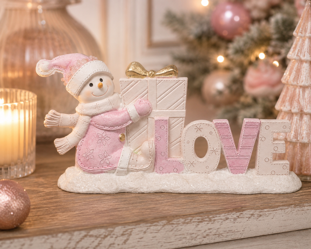 Pink snowman "LOVE" ornament holding a gift with a gold bow, styled on a mantel with a lit candle and a pink Christmas tree in the background.
