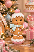 Pastel gingerbread girl figurine displayed among Christmas tree ornaments and gingerbread house décor, holding a pink swirl lollipop, styled on a wooden surface with pink and blue baubles