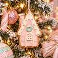 Pink gingerbread house hanging ornament with white icing roof and mint green wreath on a frosted Christmas tree with pink baubles and fairy lights