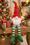 Red knit Christmas tree gnome sitting on a decorated mantel with arms outstretched and legs dangling, surrounded by wrapped gifts, ornaments and a lit Christmas tree
