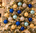 Blue, gold and silver Christmas bauble set displayed on a snow-flocked Christmas tree with warm fairy lights and gold ribbon, showing full colour mix in situ