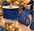 Sapphire blue glitter mesh wired ribbon roll unspooling beside a lit Christmas tree, blue and silver baubles and a wrapped gift with a navy velvet bow