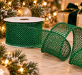 Emerald glitter mesh wired ribbon roll unspooling beside a lit Christmas tree, gold baubles and a wrapped gift with a dark green velvet bow