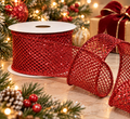 Ruby red glitter mesh wired ribbon roll unspooling beside a lit Christmas tree, red baubles, pinecones and a wrapped gift with a red velvet bow