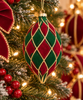 Red and green velvet harlequin Christmas bauble with gold cord diamond pattern hanging on a snowy lit Christmas tree, surrounded by red berries and red velvet ribbon