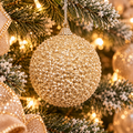 Champagne gold glitter bead Christmas bauble hanging on a snowy lit Christmas tree, surrounded by glittering gold ribbon and warm fairy lights