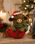 Highland cow Christmas figurine wearing a Santa hat and green scarf, sitting inside a red holly-patterned mug on a rustic wooden table with candles and a Christmas tree in the background