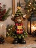 Highland cow Christmas figurine dressed as a pine tree with a gold star and holding a wrapped gift, displayed on a wooden table with a vase, lantern and Christmas tree in the background