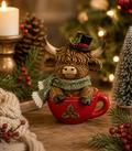 Highland cow Christmas figurine wearing a black top hat with holly trim and a glittery green scarf, sitting inside a red mug on a wooden table with a candle, pinecones and a small Christmas tree in the background