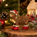 Highland cow Christmas ornament wearing red Santa boots, a Santa hat and a holly scarf, hanging on a gold cord in front of a Christmas tree with a red bauble and small light-up house in the background