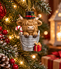 Highland cow in a bucket Christmas ornament wearing a black top hat, holding a candy cane, hanging on a snowy Christmas tree with a red bauble and gift boxes in the background