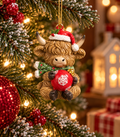 Highland cow Christmas ornament wearing a Santa hat and green scarf, holding a red snowflake bauble, hanging on a snowy Christmas tree with a red bauble and lit houses in the background