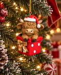Highland cow Christmas ornament wearing a Santa hat, popping out of a red gift box with a gold bow, hanging on a snowy Christmas tree with red ribbon and lights in the background