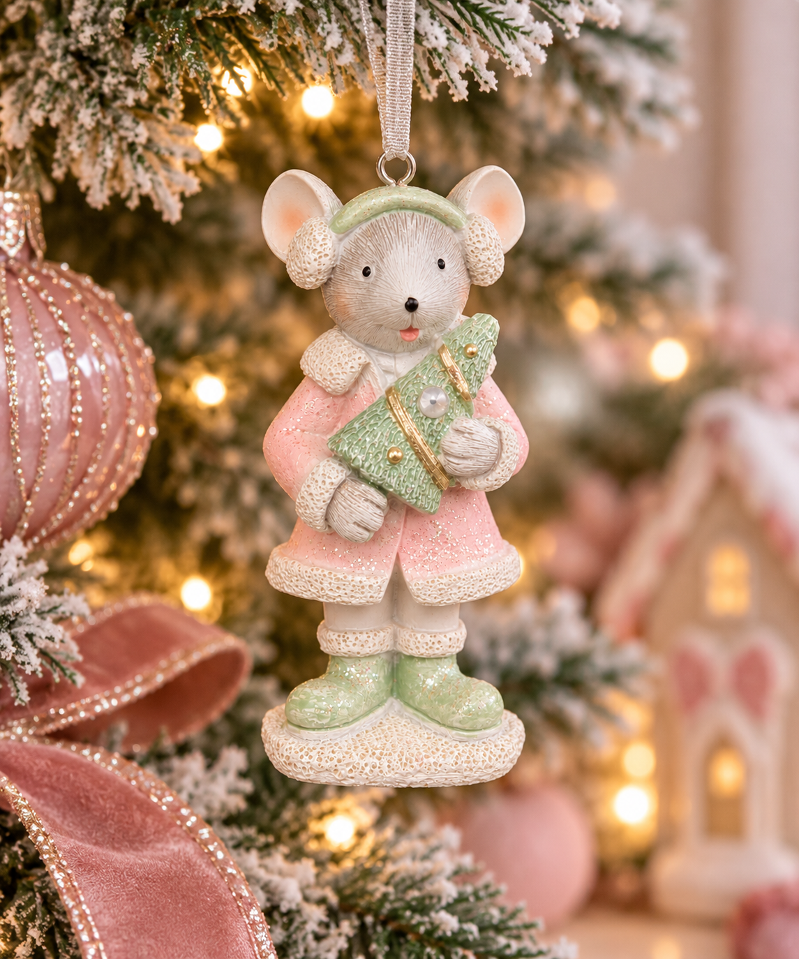 Pink coat mouse Christmas ornament hanging on a flocked Christmas tree, holding a glittered mini tree, with pink ribbon and gingerbread house display in the background