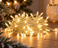 240 LED warm white fairy lights coiled and glowing on a wooden table beside a flocked Christmas tree, pinecone and glittered candle holder