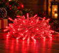 240 red LED fairy lights coiled and glowing on a wooden table beside a wrapped gift, pinecone and red baubles, with a lit lantern in the background
